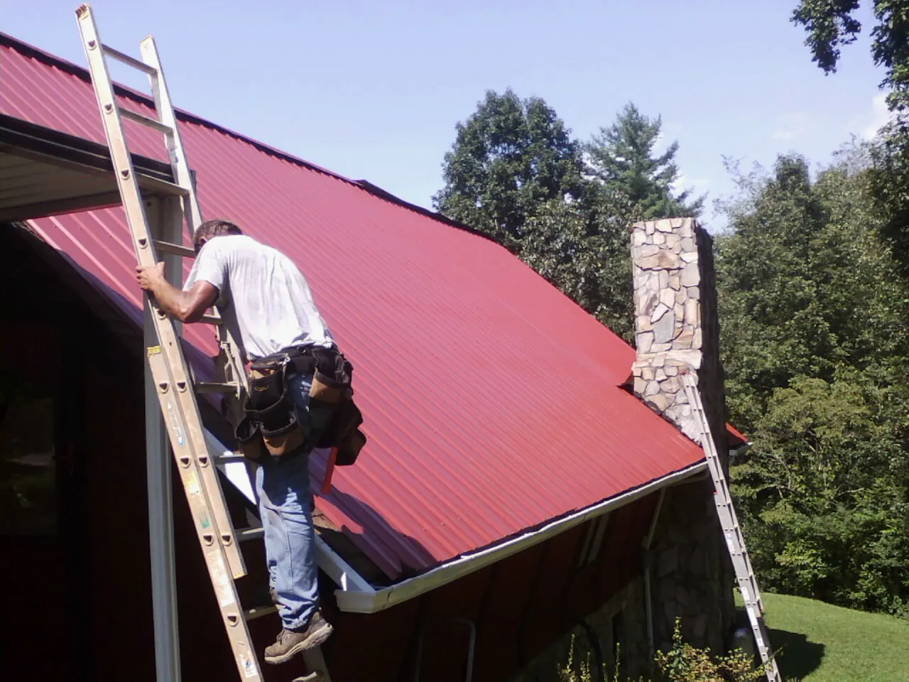 Roofer on a ladder beside a red metal roof and stone chimney — Soffit & Fascia Repair by Roofing Brothers in Raymond